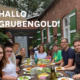 Das Team von Grubengold sitzt an einer langen Tafel voller Essen und Trinken. Im Hintergrund ein altes Backsteinhaus und Sonnenschirm.